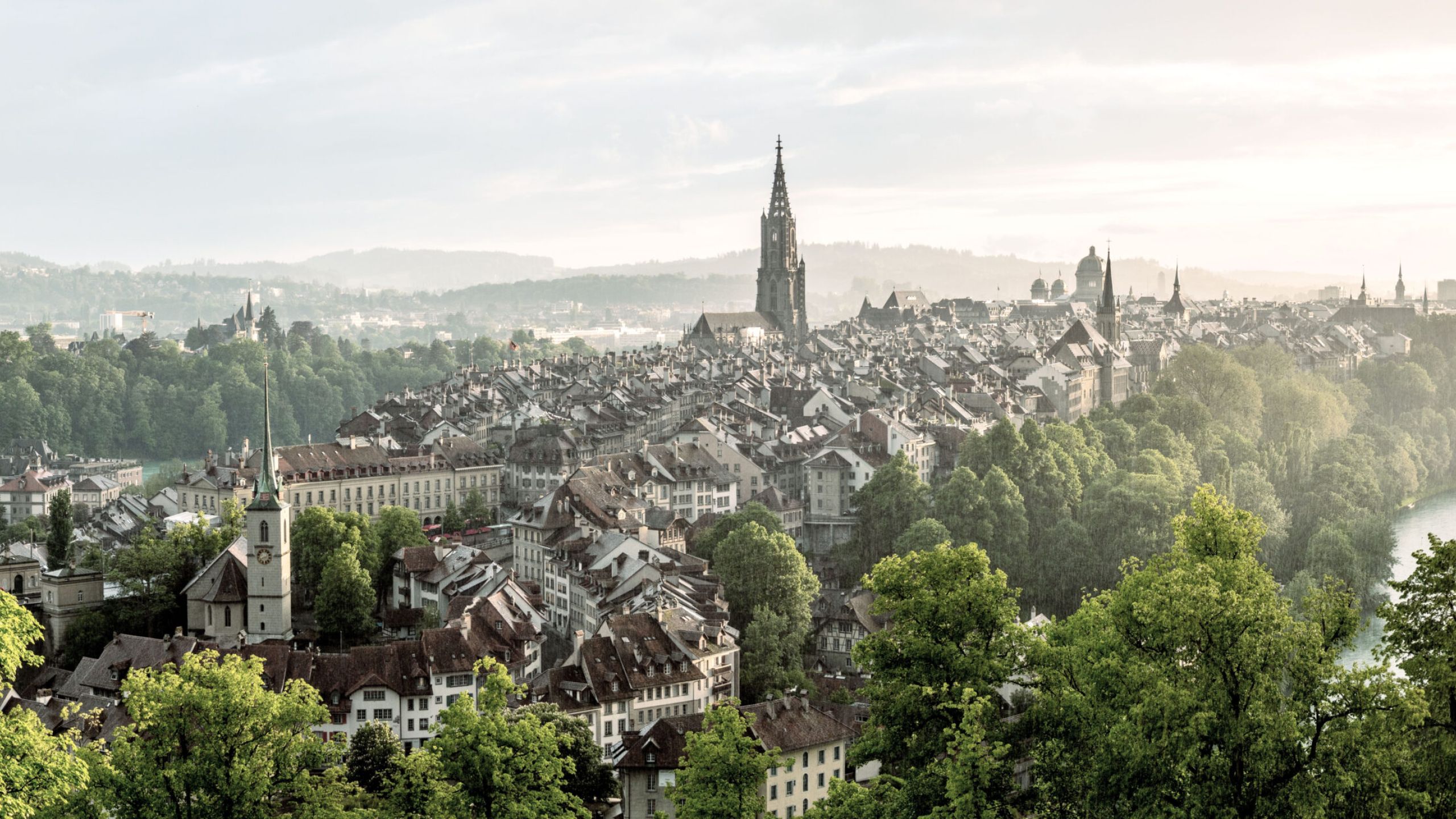 Bild der Stadt Bern von oben. Man sieht viele grüne Bäume und die Sonne die im Hintergrund untergeht.